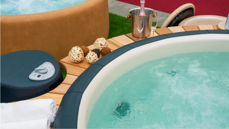 Zbliżenie na okrągłe jacuzzi z podgrzewaną wodą, obok leżą ręczniki, dekoracyjne kule i stoją kieliszki do szampana z butelką w chłodziarce